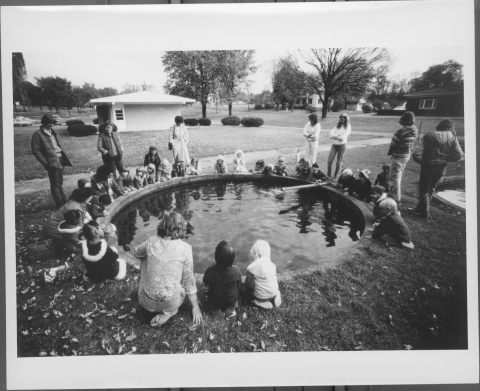 About 24 young children and about 9 adults gathered around a mid-size circular fisheries pool; the children are sitting around the perimeter of the pool, as are a couple of the adults - the rest of the adults are standing. Most of the children are dressed in coats. Another fisheries pool is just visible on the far right. A small building is visible in the background, as is a parking area on the far left. Two builds (resembling private residences) are in the background on the right.