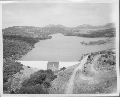 Large dam in the foreground, with a large lake beyond it. A few dirt roads are visible on the bottom right. Trees and heavy vegetation are looked in the small valley in front of the dam, and on the bank on the left of the lake. Several mountain ridges rise in the distant background.