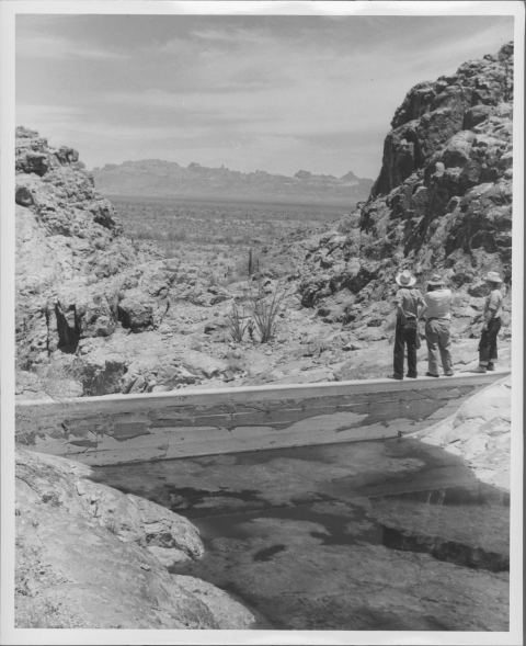 small dam in the foreground, with a body of water before it. Three men standing on the dam on the far right; the men are dressed for the field, including broadbrimmed hats. Beyond the dam is a small valley, with natural rock walls rising on either side. Beyond the valley is a flat desert, with a mountain ridge visible in the far distance. Being a desert, vegetation is small and sparse.