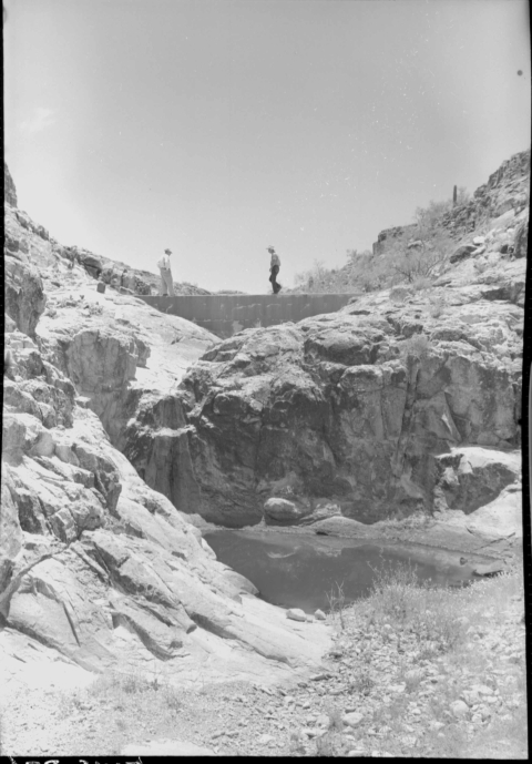 A large pool of water in the center, partially obscured to the camera by a sloping natural rock wall on the left. Another natural rock wall (perhaps part of the aforementioned wall) rises behind the pool. Atop the wall (or at least visible just beyond it) is the top of a medium-sized dam; two people stand atop the dam wall. Setting is a desert environment, with low desert vegetation in the foreground, as well as growing atop the rocky ridgelines on either side on the horizon.