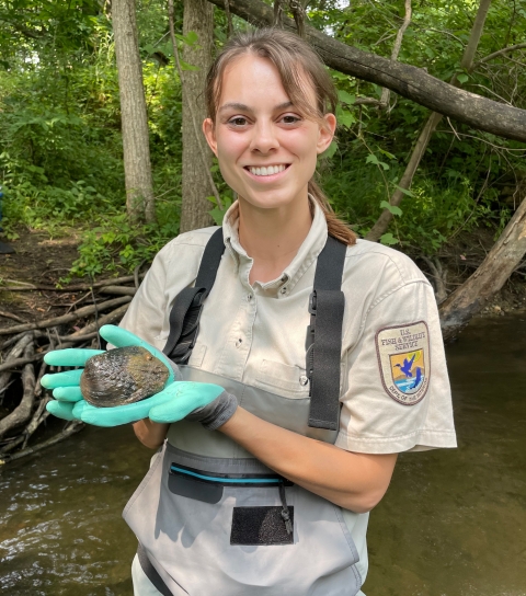 A smiling U.S. Fish and Wildlife Service biologist wading in a stream holding a mussel in her hands.