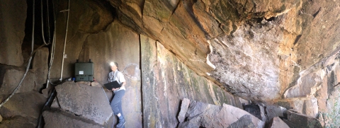a woman with a headlamp stands in the entrance of a cave holding a laptop