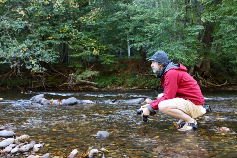 Man, holding a camera, kneeling in a shallow river