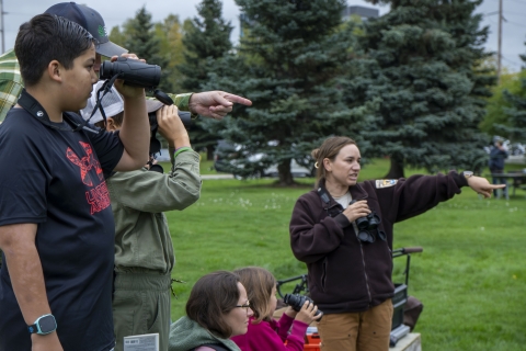 A Fish and Wildlife Biologist in a USFWS uniform looks through binoculars with a youth participant of an outreach event in a large public lawn with a parking lot in the background.