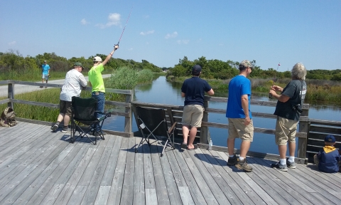 Six anglers stand on a wooden platform overlooking calm water in a small pond area. One angle is holding his fishing rod high, another appears to be actively reeling the line and a young angler is sitting on the platform.