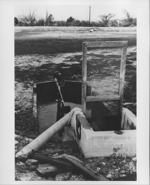 View of a person working in what appears to be a drained fisheries pond or pool; they are in part of the drained pool that features retaining walls on three sides that are about waist-high, with a smaller section facing into the aforementioned section, which features higher walls, as well as a very tall rectangular screen with a wood frame. A large pipe or tube runs into the space from the lower left corner. It appears to be winter, as a thin layer of snow is on the ground.