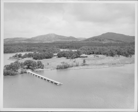 Aerial view of a small peninsula in a lake; the small peninsula features a long pier (in the foreground), several copses of trees, 3 or so small buildings/structures, and pathways. On the shore in the background is a heavily wooded area, with mountain peaks in the distance.