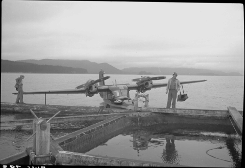 In the foreground is a large rectangular floating fish trap, consisting of a frame and crossbeams with nets (mostly underwater) attached to them. A man stands on the rearmost frame, center right, with another man on the same frame piece on the far left. Behind them is a large body of water, with a mid-size propeller-driven float plant near the trap. The distant shore can be seen in the background, complete with a ridge.