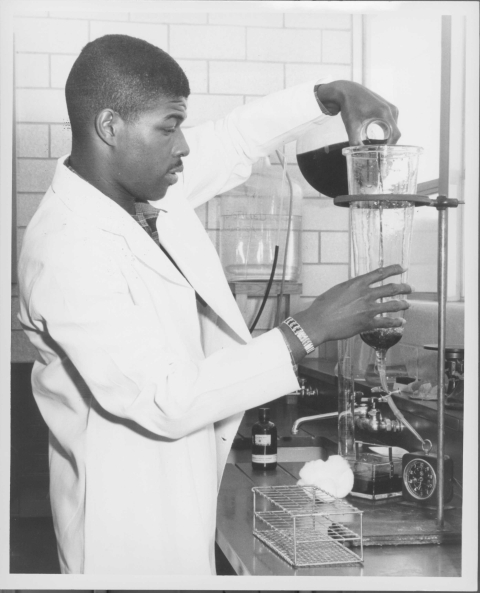 Close-up of a man, dressed in a white lab coat, in the act of pouring a liquid from a glass lab flask into a cone-shaped glass container/funnel that has a clear tube connected to it at the bottom; the container is held in a supporting apparatus that features a large metal support rod. Behind him are other pieces of lab equipment.