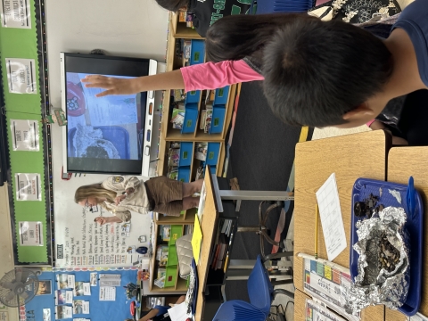 A park ranger stands in a classroom. Students are raising their hands. There is a science experiment on their desk