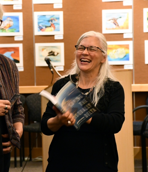 A woman with glasses and long white hair laughs as she flips through a book.