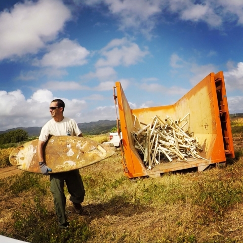 Joshua Fisher holds a beaten up surfboard near a dumpster