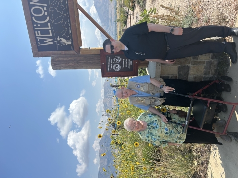 Three people stand in front of a sign. Wildflowers and mountains are in the background.