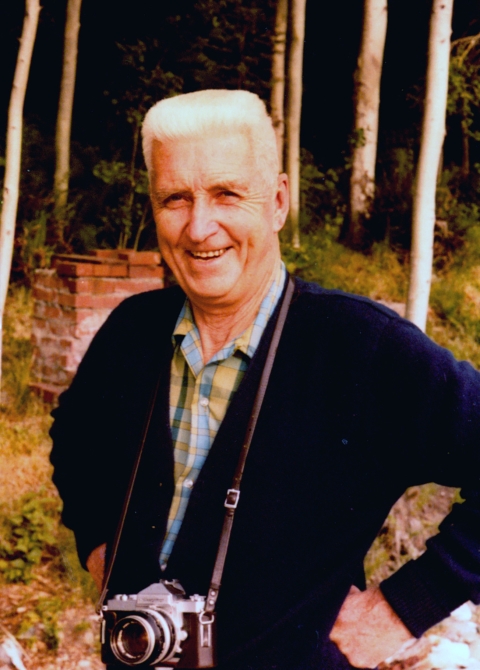 Color photo of an older man with crew-cut white hair, smiling into the camera. He wears a navy blue cardigan over a summer plaid shirt, has a single lens reflex camera around his neck on a thin leather strap, and his hands are on his hips. A brick structure and some white-barked trees at the edge of a dark wood are behind him.