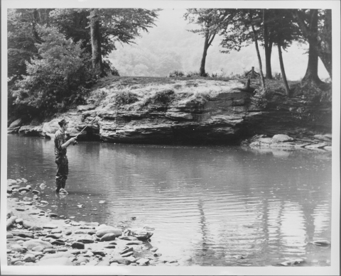 Man fishing with a rod, center left, in the shallow water near the shore of a large stream/small river. A large rocky outcropping dominates the center of the opposite shore, as well as a number of trees and large bushes. More trees are visible in the distance. The man wears rubber wading pants and a cap.