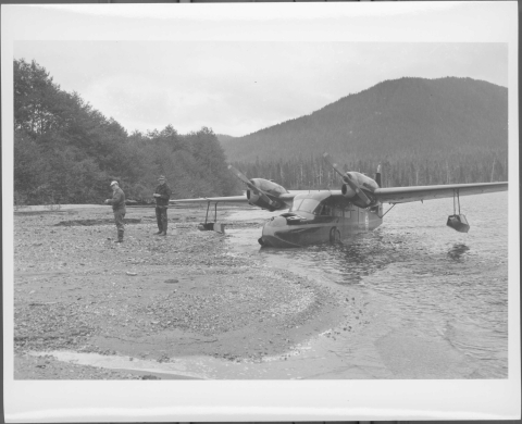 Medium-sized floatplane moored in the shallow area of a lake shore, with two men dressed for field service standing on the shore to the left of the aircraft. The shore area in view is slightly rocky. A line of trees in visible in the left background, with more trees visible on distant lake shore in the center and right background. A mountain ridge line rises in the distance on the right.