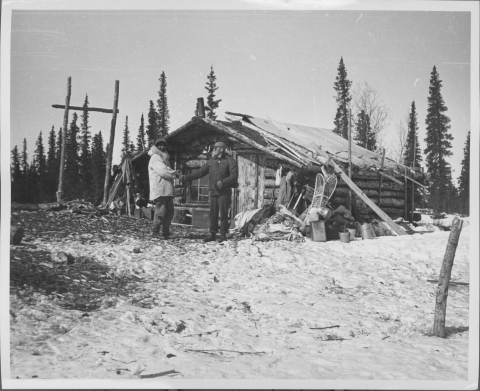 Two men standing in front of a small log cabin/dwelling, shaking hands. Both are dressed for cold weather. Snow covers the ground, mainly in the foreground and on the right. In background beyond the cabin is a line of evergreen trees.