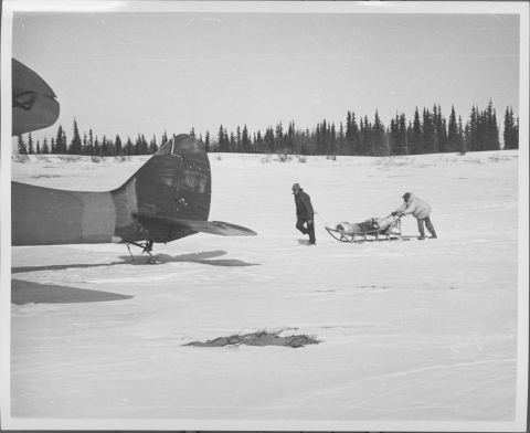 Two men in the middle distance, just right of center, moving a wooden-framed sledge through a snow-covered landscape; one man is in front, pulling the sledge, while the other man is in back, pushing it. The tail section and part of a wing tip of an airplane protrudes in to the frame from the left. Evergreen trees line the horizon.