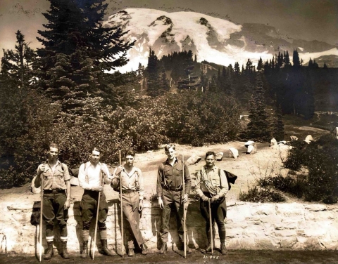 5 young men stand by a low stone wall, dressed in hiking boots and holding staffs, with Mount Rainier filling the sky behind them and a view of trees and shrubs on a slope.