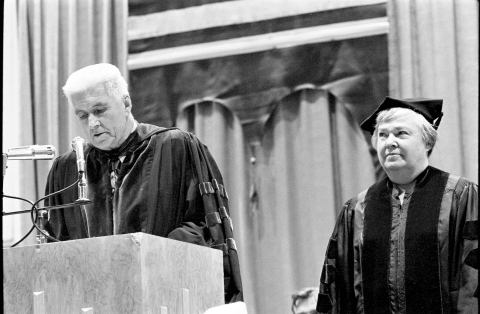 Black and white photo of a man wearing college robes speaking into microphones at a podium, with a woman in similar robes and a graduate's hat behind him.