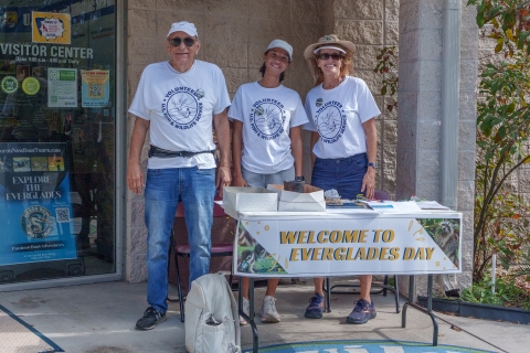 Three volunteers greet people in front of the visitor center. A sign on the table says "Welcome to Everglades Day"