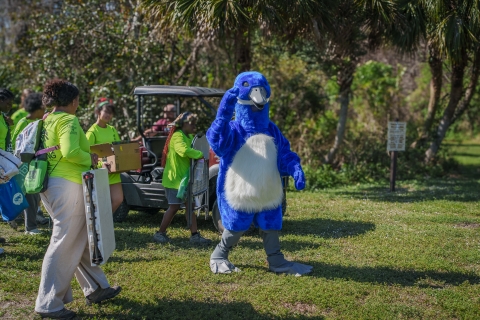 A blue goose mascot waves from the field. A group of people wearing green shirts is carrying materials.