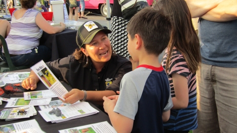 Woman wearing USFWS logo talking with children at a table about pollinators, with a pollinator outreach kit laid out on the table.