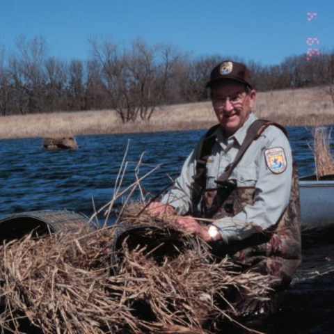 Biologist wearing waders and sitting in a boat on the water fixing a grass/straw hen house that sits on top of the water.