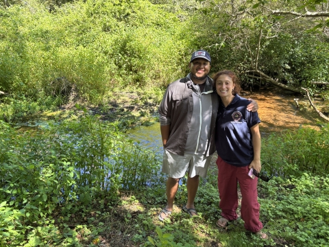 a man wearing a gray-and-white shirt, white shorts, and a blue ball cap stands beside a woman wearing a blue shirt and red pants on a plant-covered river bank, with the river behind them