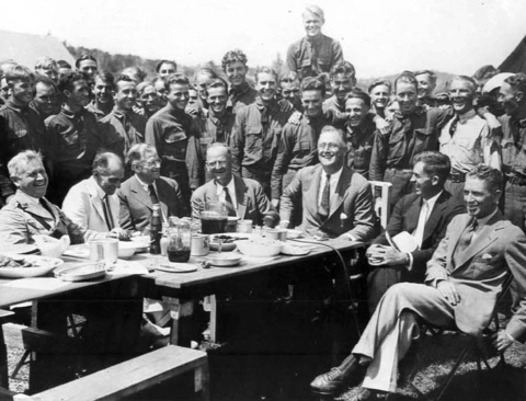 b&w photo of men in suits at table outside with CCC members behind