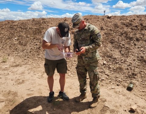 a fish and wildlife service biologist and military personnel dressed in fatigues looks down at a container with a lizard