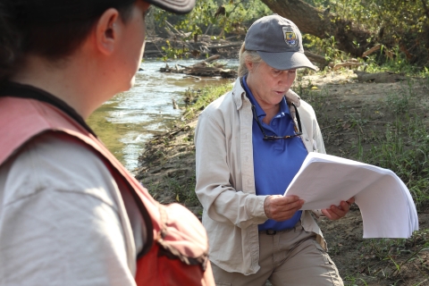 Two people standing beside a river, one looking at papers in her hand