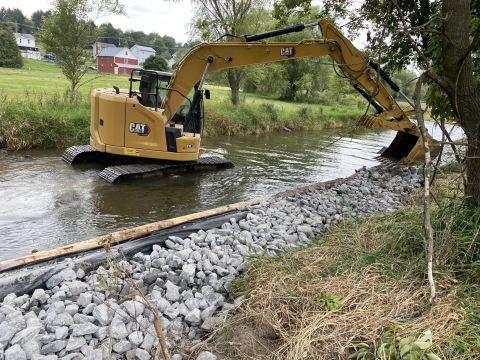 Excavator in a stream moving boulders to stabilize the bank. There are boulders along the right side of the bank that the excavator moved.