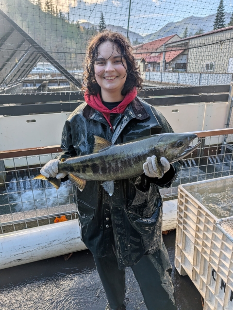 WaterCorps Service Member holding an adult chum salmon. Fish hatchery and skyline in the background.