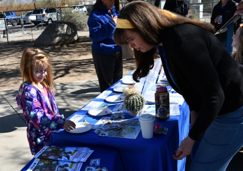 a child stands near a table with bird-related activities while a biologist talks with her