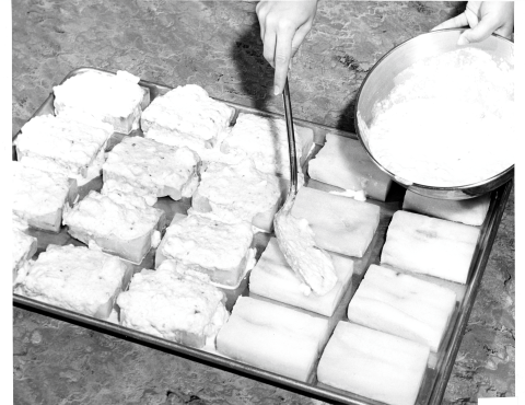 person (out of frame, with the exception of their hands) spooning sauce from a bowl on to 20 fish fillets, arranged on a baking sheet