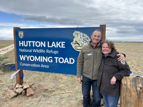 Two people posing for a photo outside in front of a sign that reads, “Hutton Lake National Wildlife Refuge Wyoming Toad Conservation Area”. The sign is dark blue and has the U.S. Fish and Wildlife Service logo on it and a picture of the Wyoming Toad
