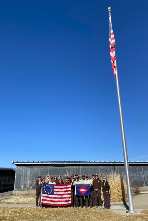 US flag flies, while staff on ground hold Betsy Ross flag, fish pennant
