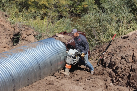 Person using a large motorized hand tool to install a culvert in the bare ground. There is some green vegetation in the background.