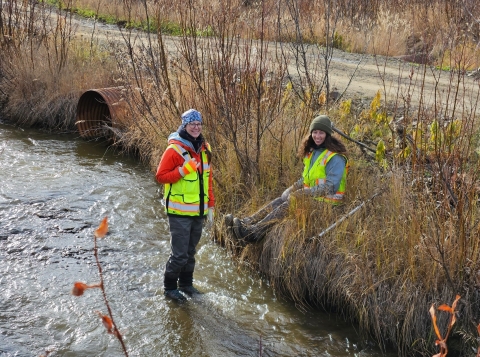 Two people wearing waders and reflective safety clothing are working at a stream crossing. One person stands in the water, while the other sits on a vegetated bank. A road with a culvert running underneath it is visible in the background.