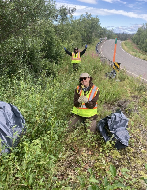 Two people in safety vest smile at camera holding up invasive toadflax on roadside. Trash bags are visible in the foreground from handpulling the invasive plant.