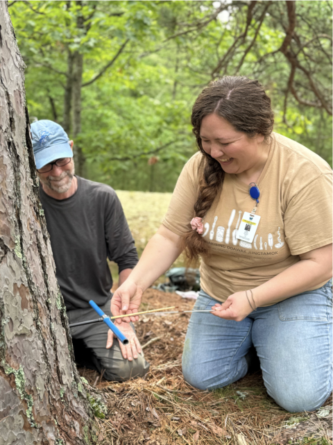Person takes core sample of tree in the foreground while another person supports the work in a training capacity