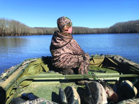 A child in camouflage on a boat with decoys near their feet looks out at the water.