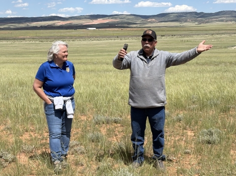 Two people stand in a grassy field outdoors; one person speaks with hands outstretched while the other watches and listens.