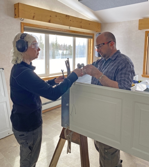 Two people indoors wearing hearing protection and safety glasses, holding a white garage door panel propped up on a wooden stand and hand tools.