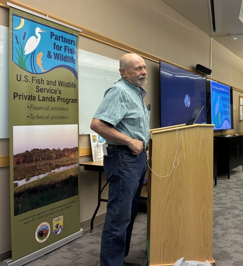 A man standing behind a podium with talking with a vertical banner in the background that has the Partners for Fish and Wildlife Program signature on it and "U.S. Fish and Wildlife Service's Private Lands Program".