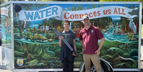 Service employee and youth event participant posing in front of an event trailer. The trailer is decorated with a below and above surface nature scene with rivers, habitat, fish, amphibians, birds, people, and mountains.