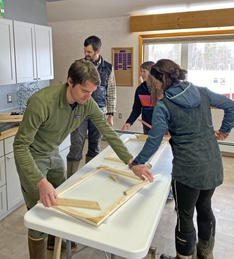 Four people indoors constructing two separate wooden frames on a white table