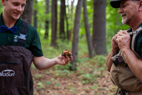  A man wearing hip waders and a blue bowtie holds a small turtle out to another man in waders. They are standing in a wooded area.