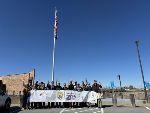 Group of people standing outdoors holding a USFWS-America 250 banner beneath a flagpole; flags flat
